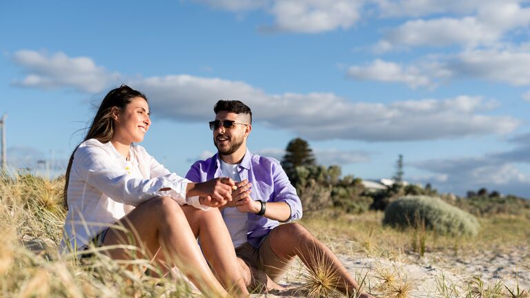 Two people sitting on the sand at the beach. One is wearing a white button up long sleeve and the other is wearing a purple long sleeve button up with sunglasses.