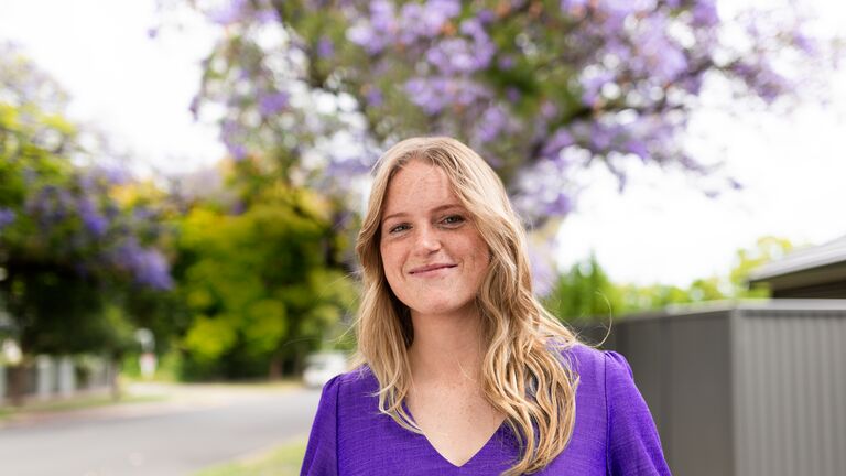 Woman wearing a purple long sleeved top standing in front of a jacaranda tree