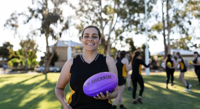 Person wearing a plain black football guernsey smiling at the camera while holding a purple football in their hands.