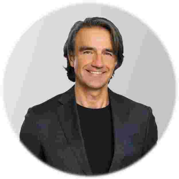 Headshot of a smiling man with graying hair, wearing a dark suit jacket over a black shirt.