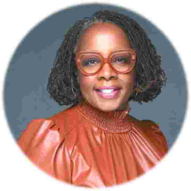 A smiling woman with dreadlocks, wearing large orange glasses and a rust-colored top.