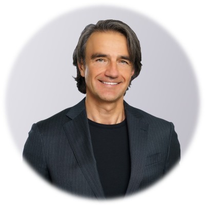 A smiling man with dark, medium-length hair wearing a dark suit jacket over a black t-shirt.