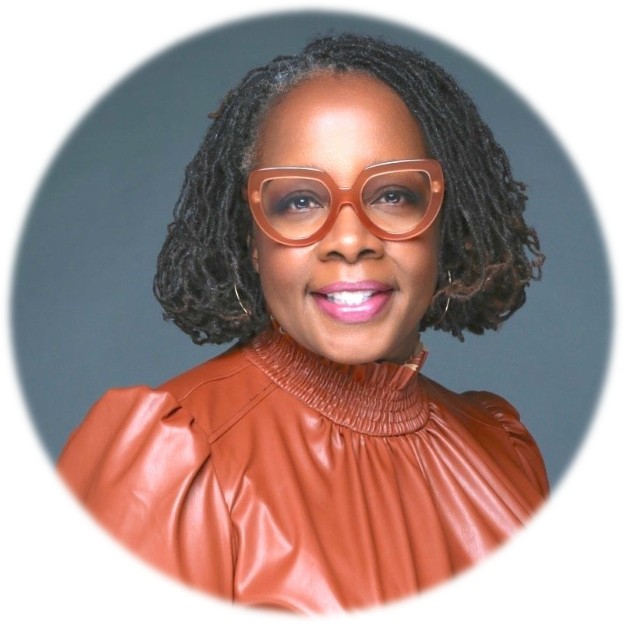 A smiling woman with dreadlocks and large orange glasses, wearing a brown leather-look top.