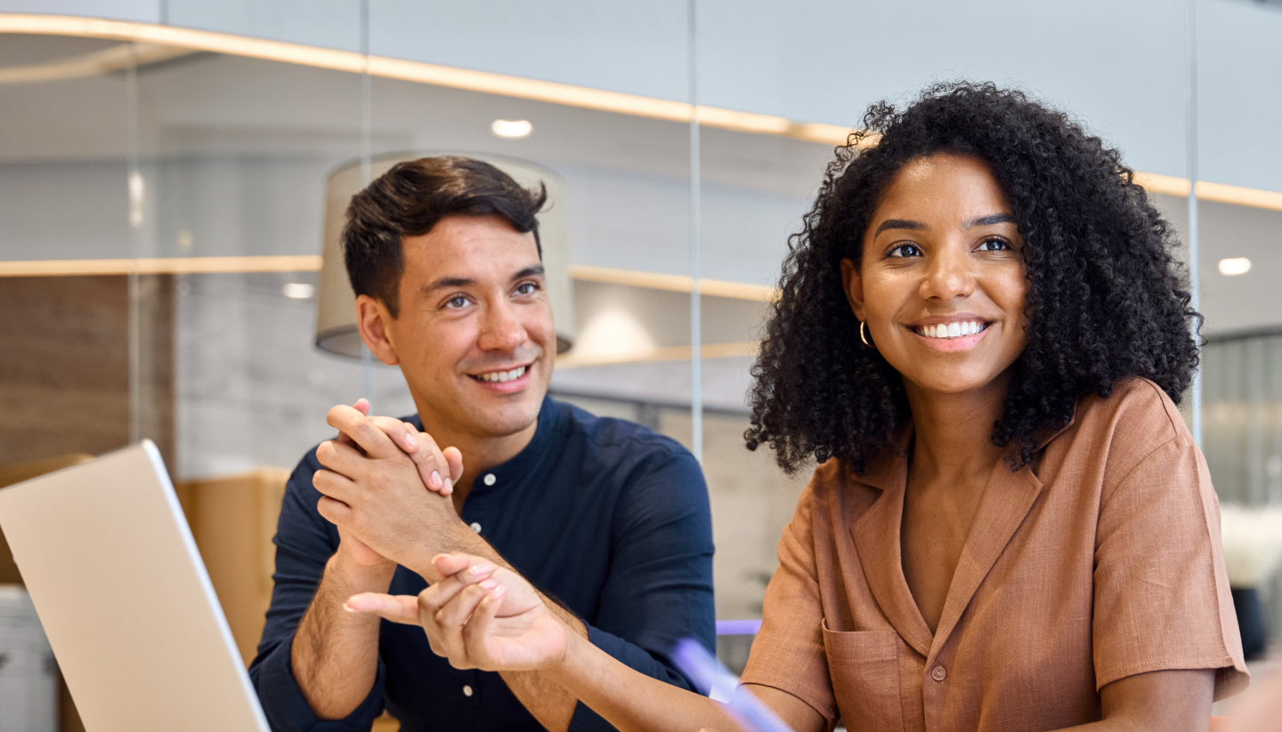 Two office employees smile as they listen to a co-worker in a meeting.