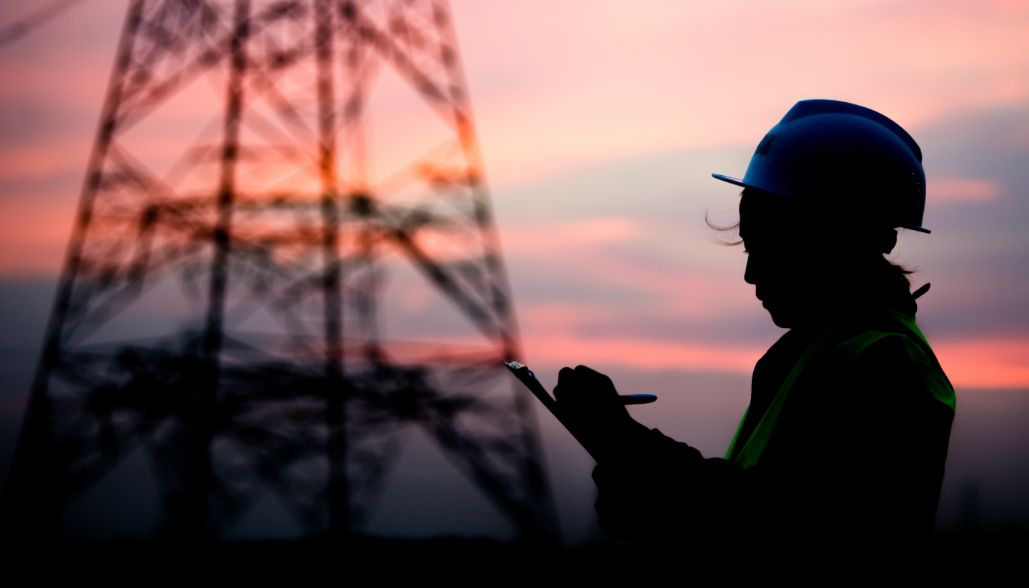 A silhouetted construction worker writes on a clipboard at a project site.