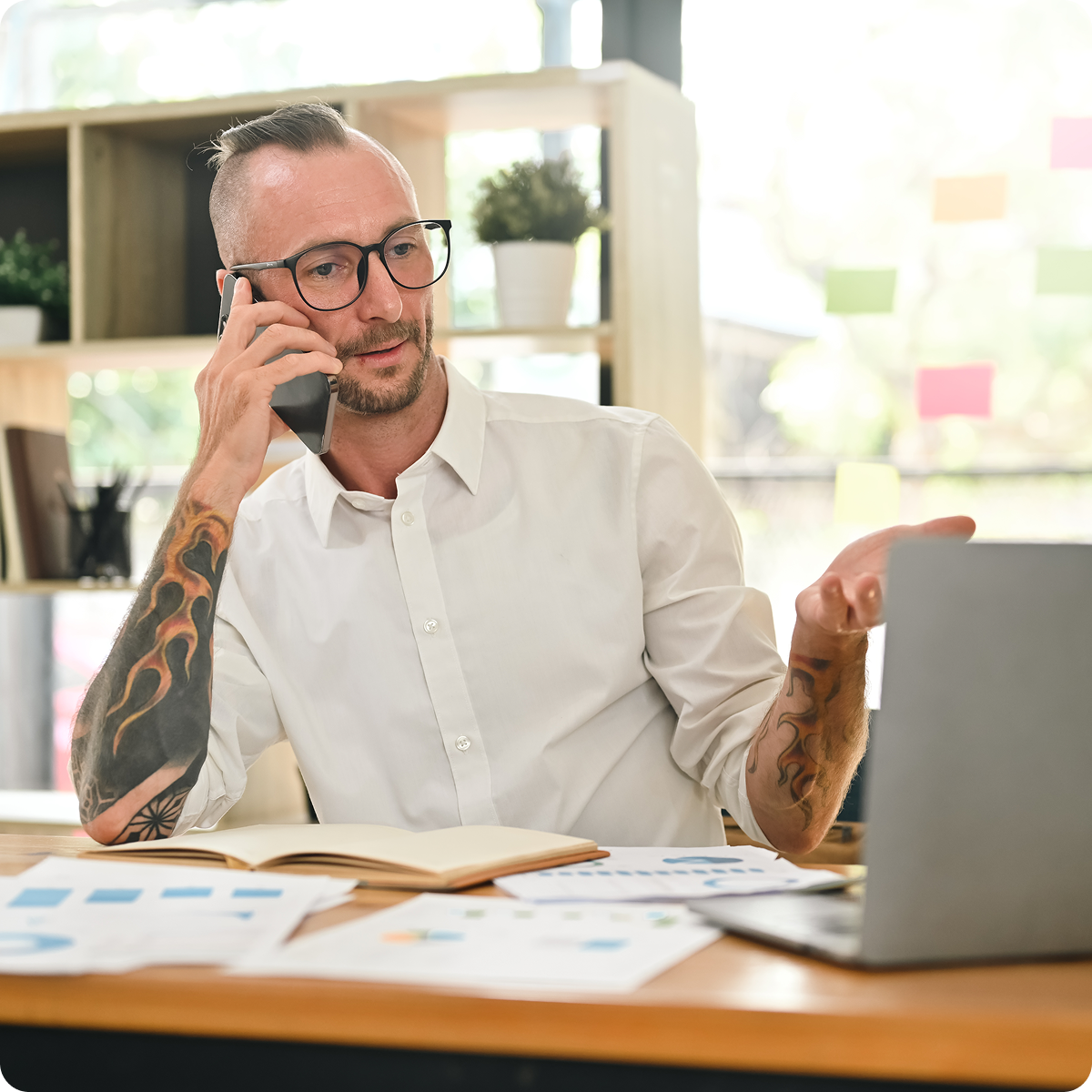 Construction office worker sitting at a desk with multiple printouts in front of him while looking at a laptop screen and talking on the phone