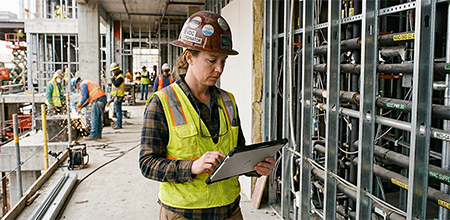 A specialty contractor in a hard hat and safety vest uses a tablet to inspect exposed wall pipes at a construction site.