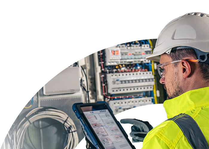 Ein Techniker mit Schutzhelm und gelber Warnweste überprüft Daten auf einem Tablet, während er in der Nähe von Schalttafeln arbeitet.
