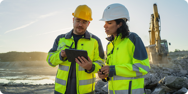 A male and female worker with neon jackets discuss over a tablet.