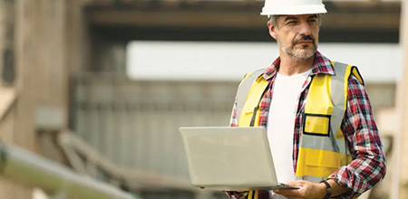 Image shows a man in a hard hat and high vis on site, holding a laptop