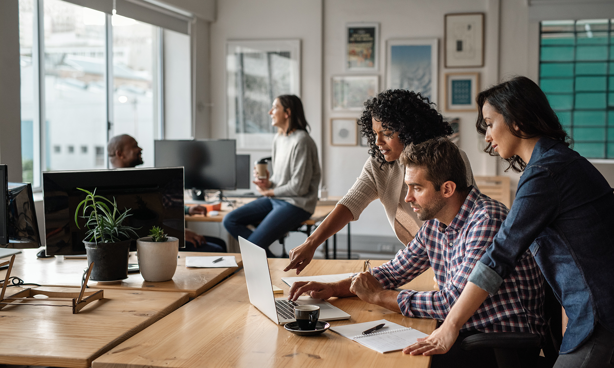 Three colleagues gathered around a laptop collaborating on a project in a modern office.