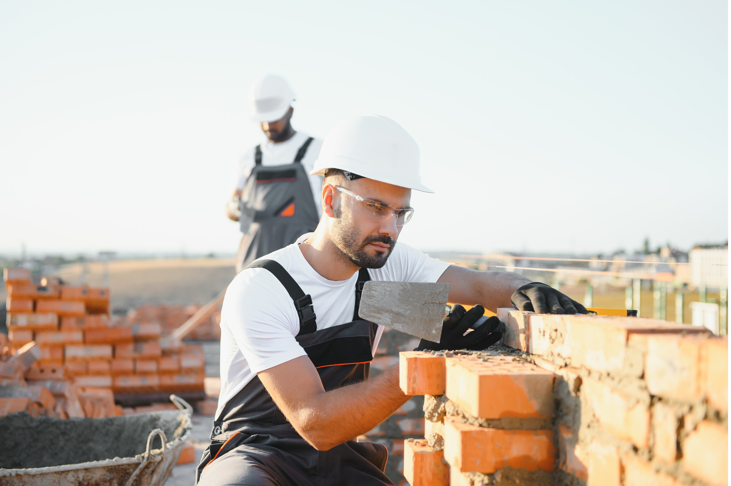 A man in a hard hat and safety glasses lays bricks while another man stands in the background looking down.
