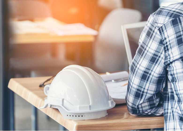Hard hat on desk next to construction worker looking at a laptop and paper documents