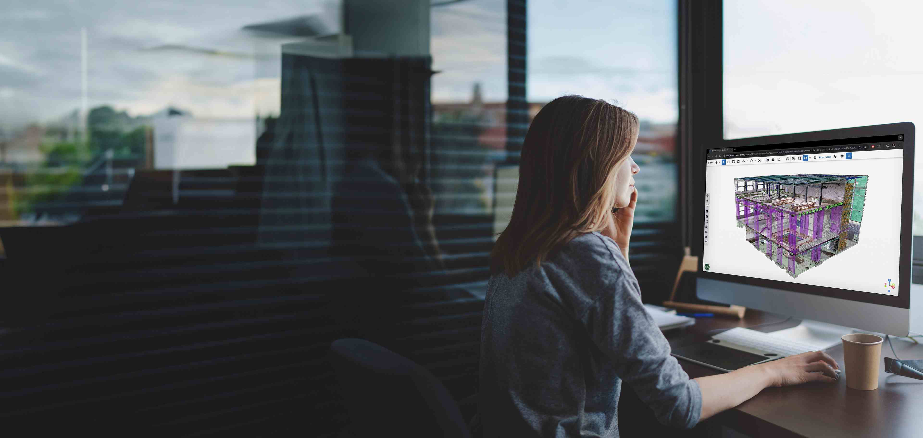 Een vrouw in een modern kantoor gebruikt Trimble Connect op een mac computer om een gedetailleerd 3D-constructiemodel van een gebouw te bekijken. De ruimte heeft grote ramen met uitzicht naar buiten en de sfeer is gefocust en professioneel.