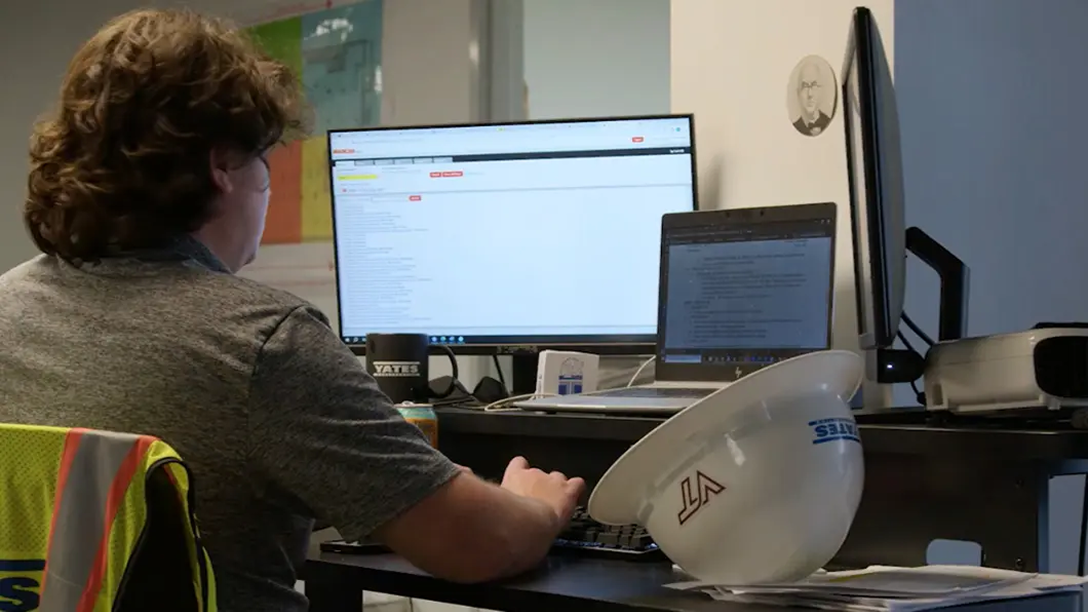 Project manager reviews documentation and tracking systems at dual monitors in office setting