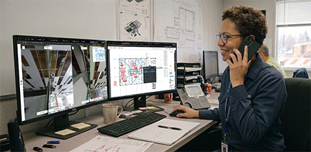 A professional on a phone call in an office, looking at dual monitors displaying construction site 360-degree views and floor plans.