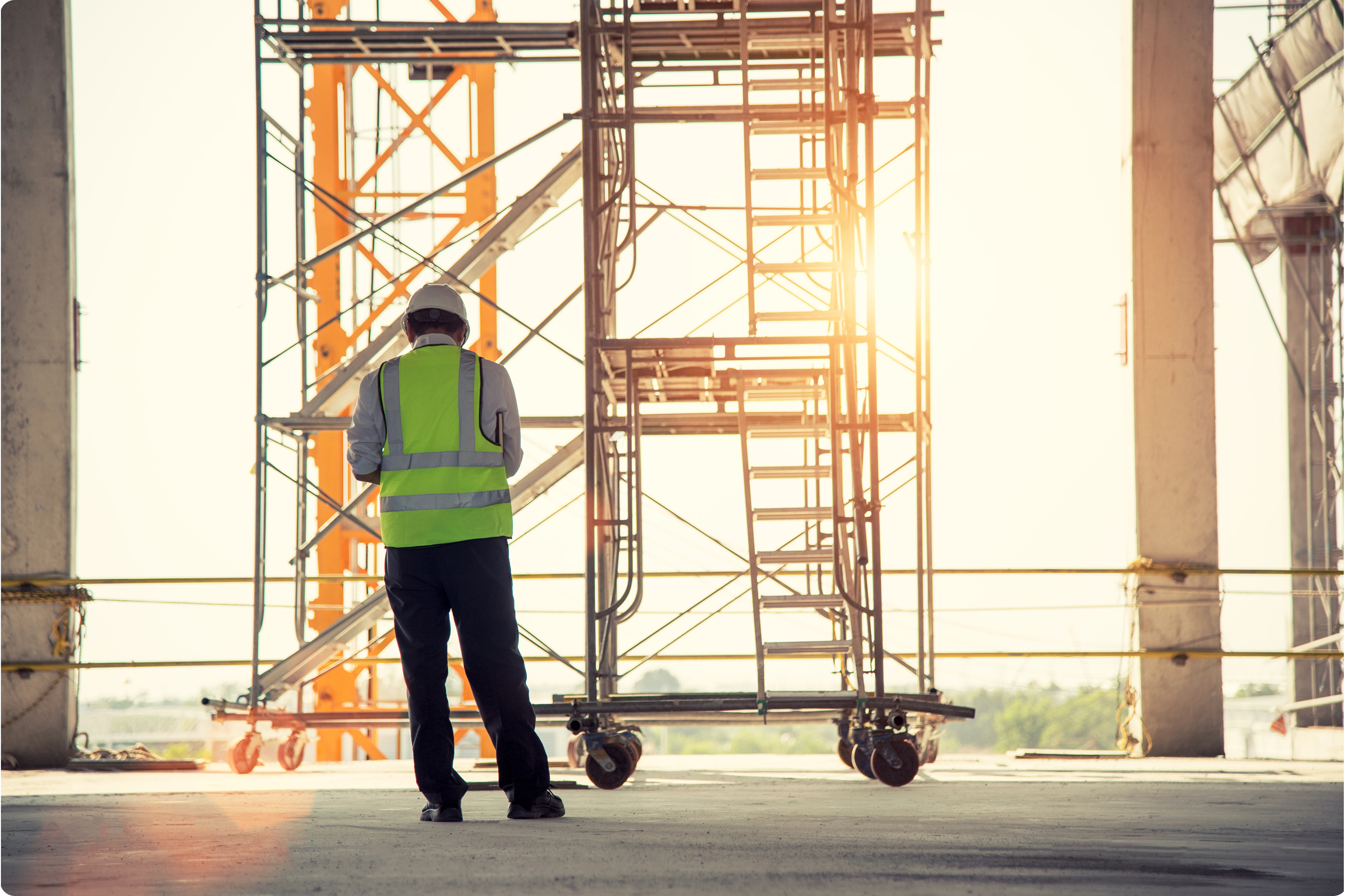 A man in a safety vest and hard hat faces away from the camera and looks down while sun shines over the site.