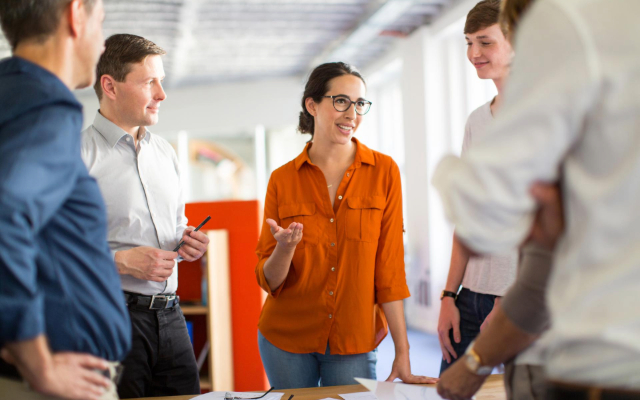 Five colleagues engaged in a vibrant conversation, looking at papers on a desk in a modern work environment.