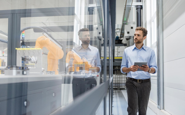 Businessman in a light blue shirt holding a tablet, observing automated machinery in a high-tech manufacturing plant.