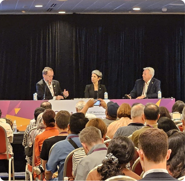 Three speakers join a conference panel before an audience, with one attendee photographing the discussion during a live session.
