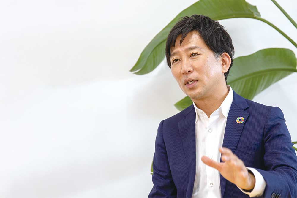 紺色のスーツを着てSDGsバッジをつけた男性が、身振り手振りを交えながら話している。