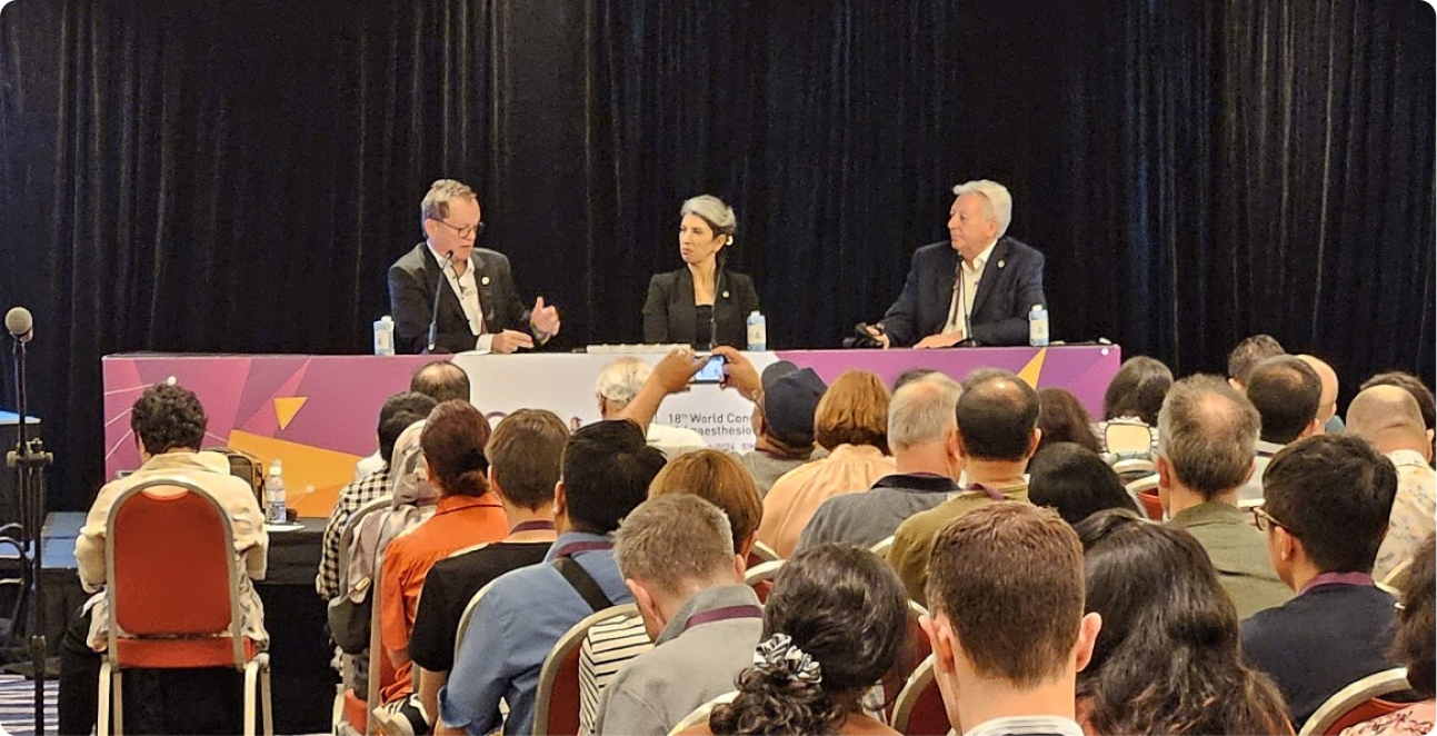 Three speakers join a conference panel before an audience, with one attendee photographing the discussion during a live session.