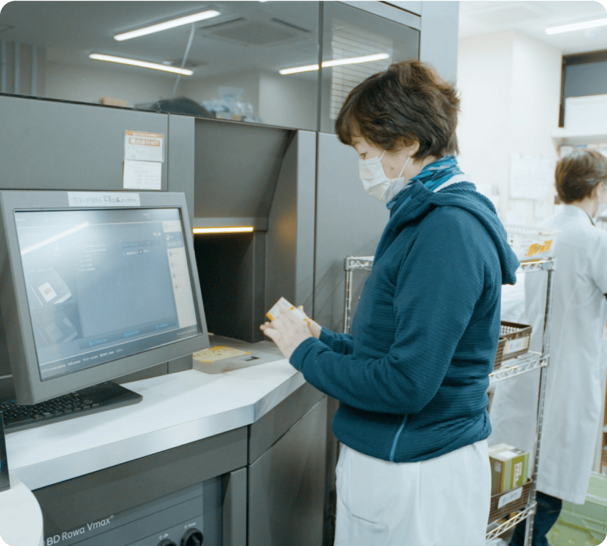 マスクをした女性が、薬局の自動調剤システムの前で薬の箱を扱っている。