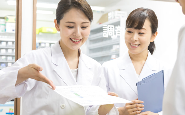 白衣を着た2人の女性が、薬局で笑顔で書類を指差して説明している。