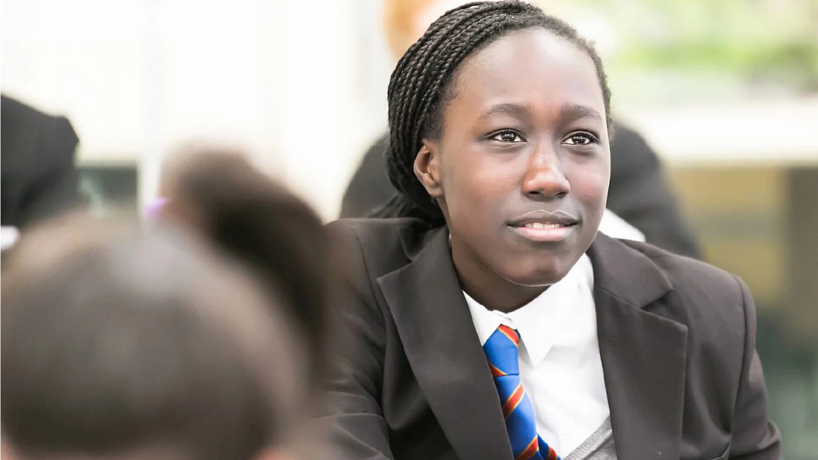 A young person with braided hair wearing a school uniform, including a black blazer and striped tie, looks attentively to the side.