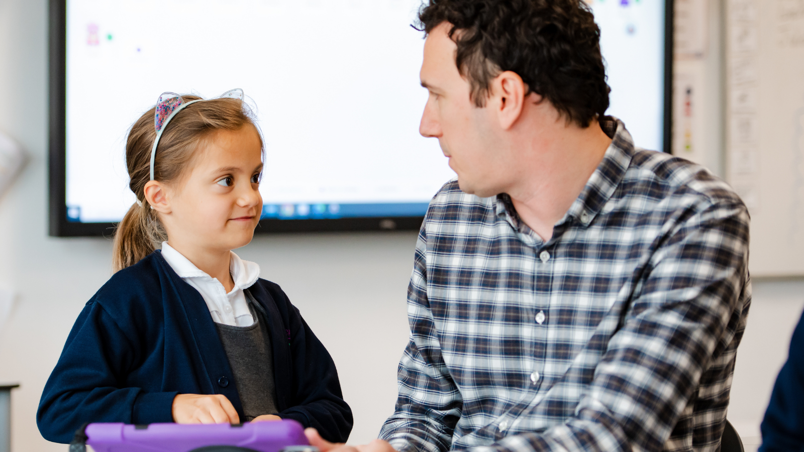 A young girl in a school uniform and a man in a plaid shirt looking at each other in a classroom.