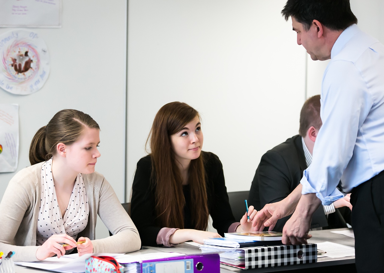 A teacher leans over a classroom table to guide students reviewing papers, binders, and writing materials.