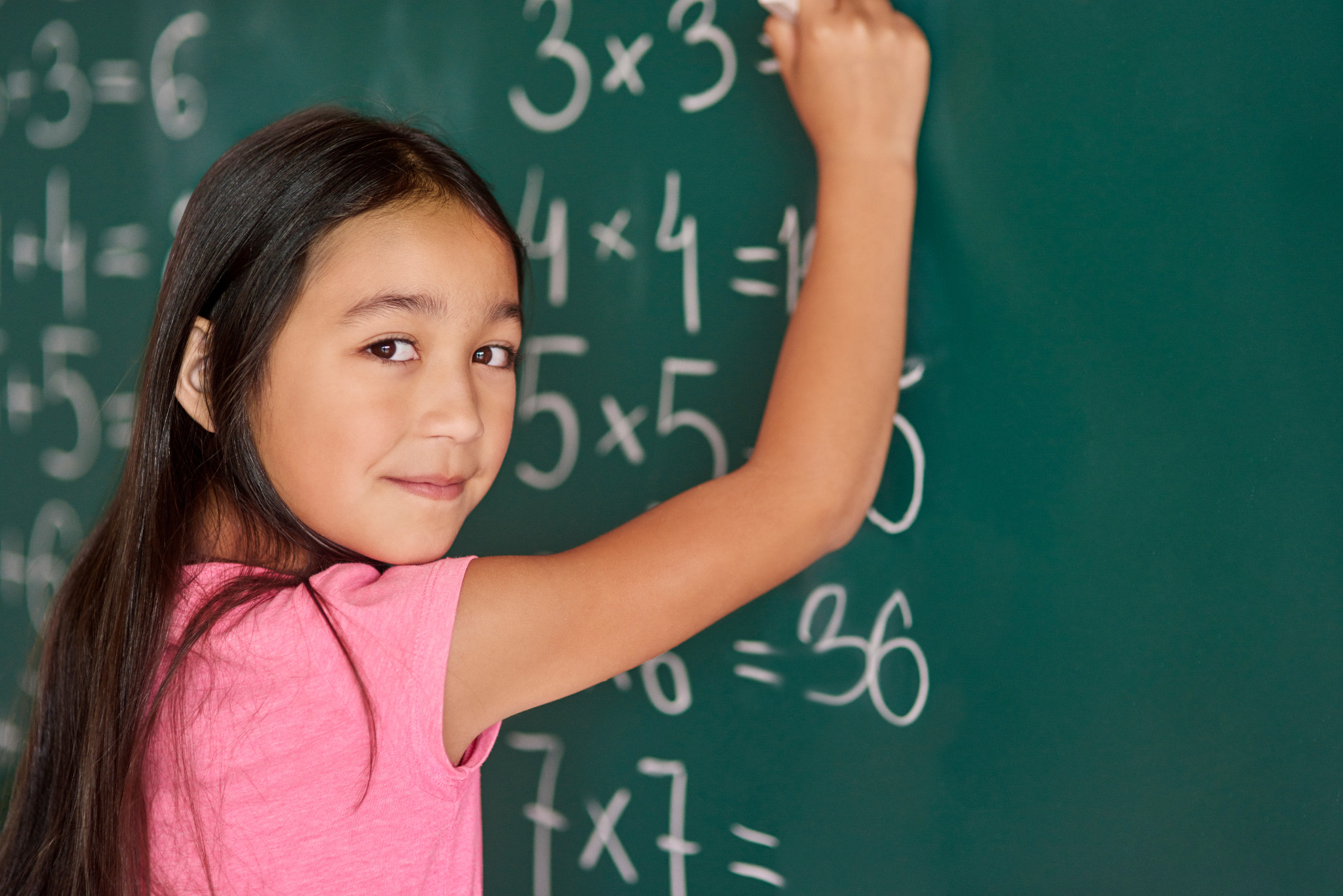 Young student in a pink shirt writes multiplication problems on a classroom chalkboard while looking at the camera.