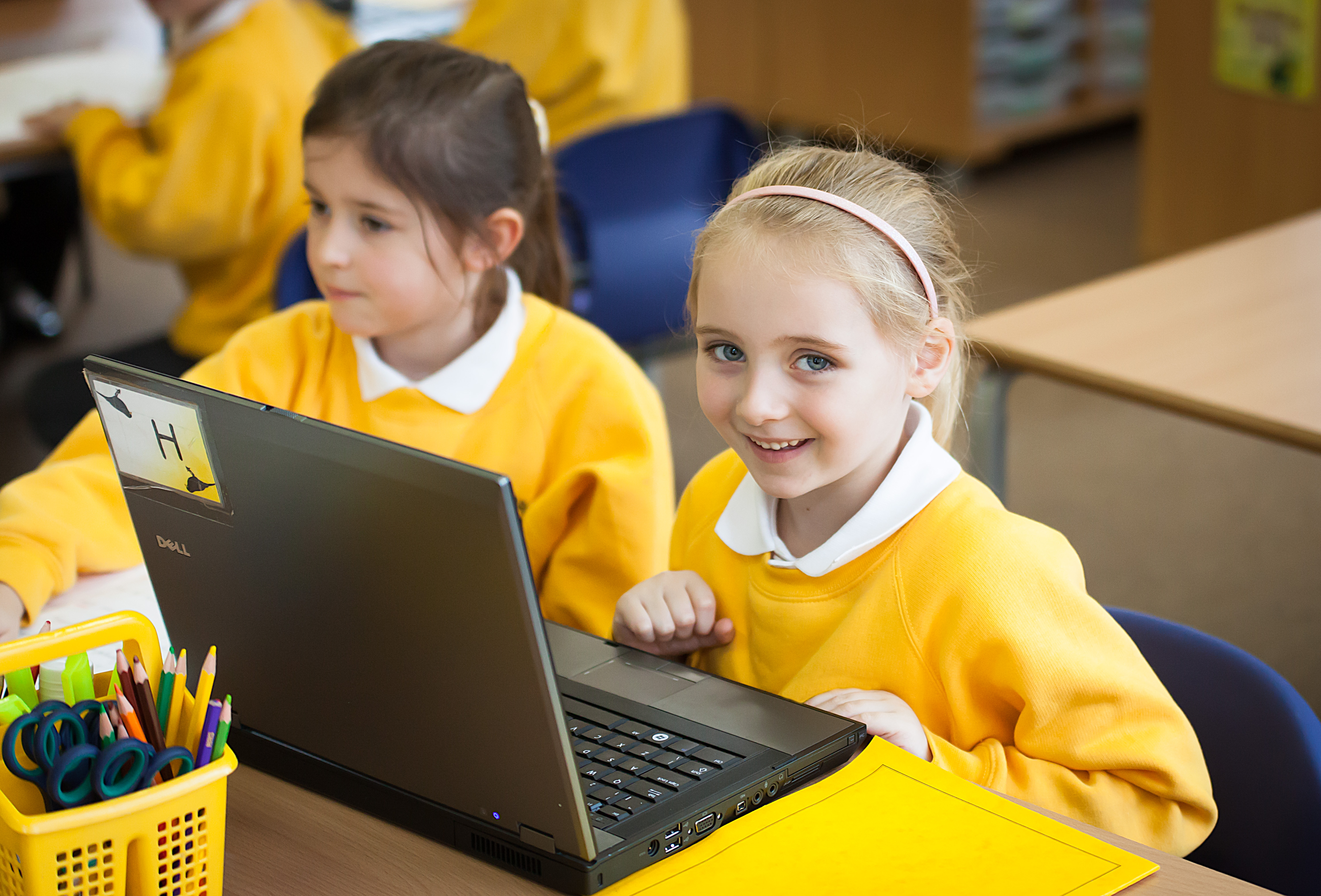 Smiling primary school student in a yellow uniform uses a laptop beside a classmate during a classroom lesson.