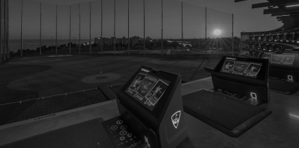 A black and white image of a Topgolf driving range at dusk, with interactive screens in the foreground and a setting sun over the water in the background.