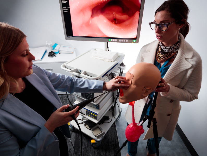 Two women practice using an endoscope on a medical mannequin, with an internal view displayed on a monitor.