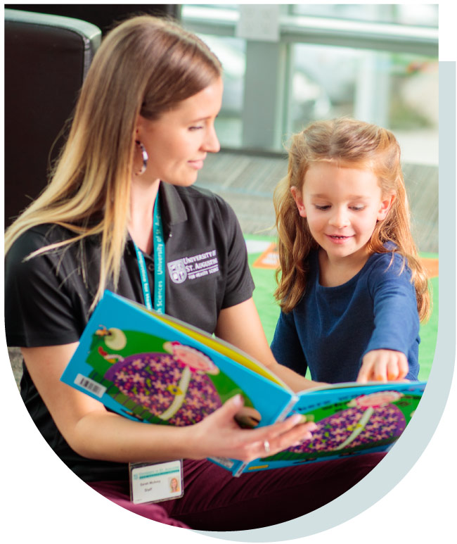 A woman and a young girl smiling as they read a colorful picture book together, with the woman wearing a University of St. Augustine polo shirt.