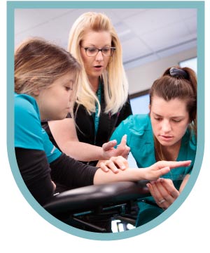 An instructor demonstrates a procedure on a student's arm to another student, all wearing teal uniforms in a learning environment.