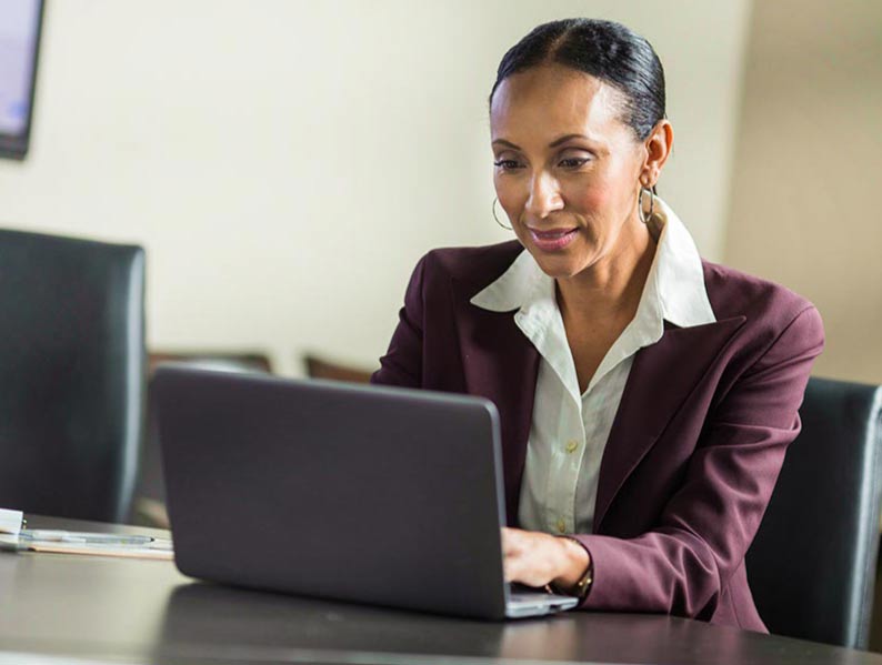 A professional woman in a maroon blazer and white shirt works on a laptop in a meeting room.