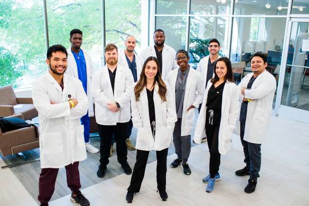 A diverse group of smiling medical students or professionals in white lab coats and scrubs, standing in an indoor setting with large windows.
