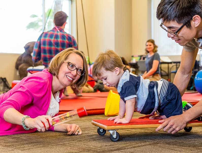A smiling woman in glasses and pink cardigan interacts with a toddler on a wheeled cart, guided by a man in a therapy or play setting.