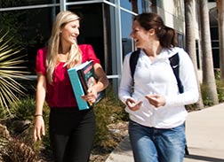 Two smiling female students walking and talking on a college campus.