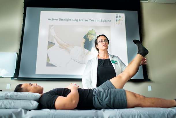 A medical professional assists a patient performing a supine straight leg raise, illustrating a physical therapy technique with a presentation slide visible.
