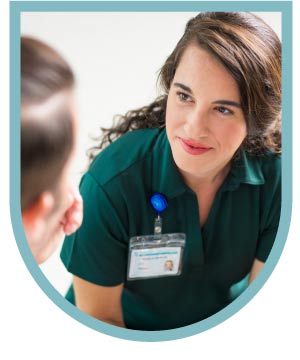 A female healthcare professional in a green uniform and ID badge smiles while looking at a patient.