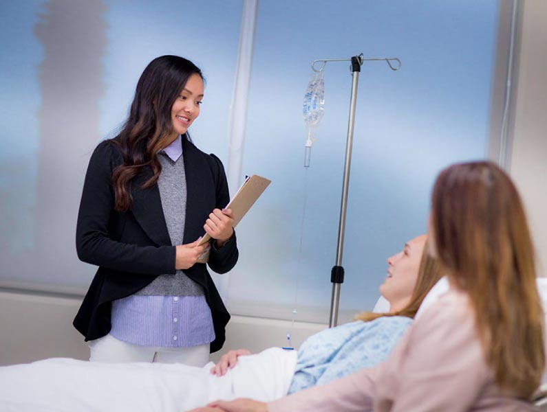 A healthcare professional with a clipboard speaks to a smiling patient in a hospital bed.