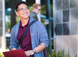 A smiling young man in glasses and a grey cardigan holds a red laptop, looking up happily while walking outside a modern building.