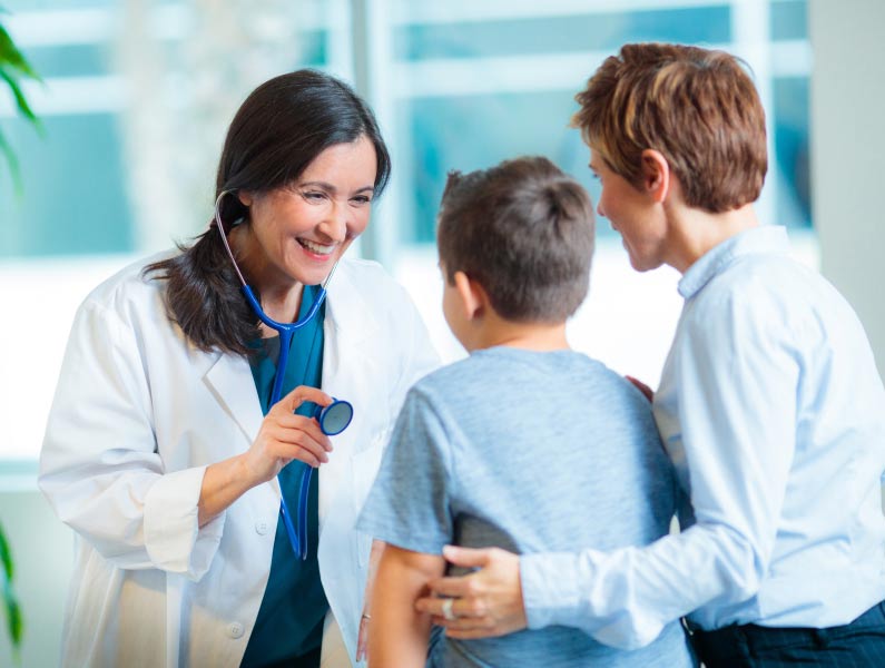 A female doctor interacts cheerfully with a child during a medical check-up, with an adult woman observing.