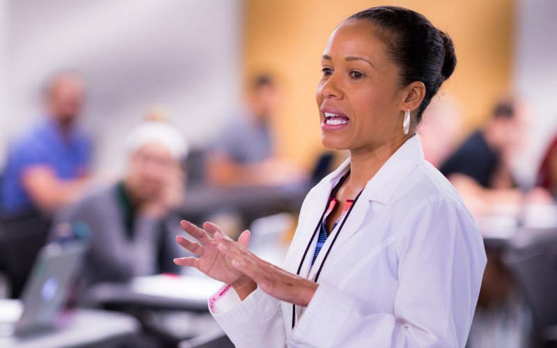A female doctor or educator, wearing a white lab coat, passionately explains something to a class.