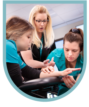 Three women in a professional setting, one instructor guiding two students in a practical demonstration.