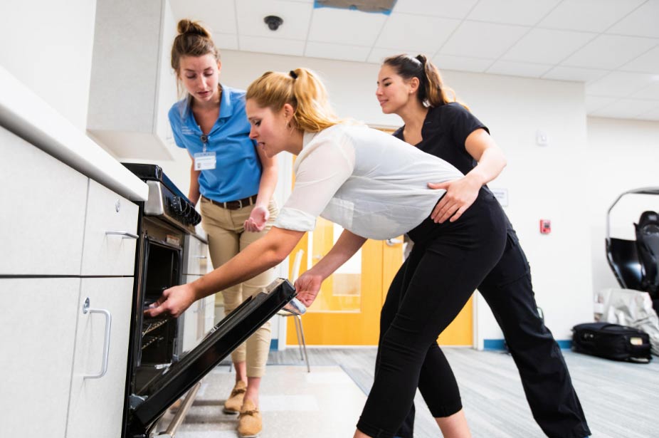 Three women in a modern kitchen, with one being assisted by another as she reaches into an open oven, demonstrating occupational therapy.