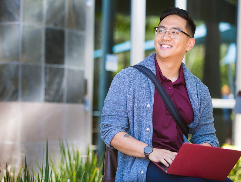 A smiling man wearing glasses and a gray cardigan works on a laptop outdoors on campus.